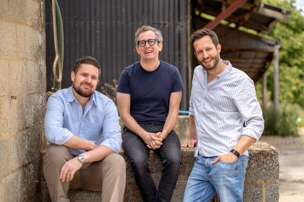 Three men smiling at the camera outdoors beside a concrete block wall, two seated and one standing and leaning on the wall, in front of a dark corrugated farm building with trees behind.